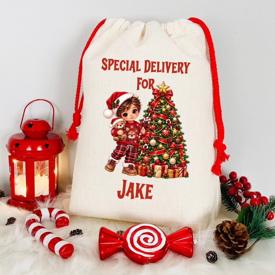 A cream-colored canvas Christmas gift bag with red drawstring cords, featuring a printed illustration of a child in a Santa hat next to a decorated Christmas tree and the text 'SPECIAL DELIVERY FOR JAKE'. The bag is styled on a white faux-fur surface with red holiday props including a lantern, candy canes, a lollipop-shaped ornament, pinecones, and berry sprigs.