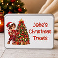 A rectangular tin with rounded corners and a silver metal border, featuring a white front label with a Christmas illustration of a child in a Santa hat next to a decorated tree and the text 'Jake's Christmas Treats' in red. The tin is photographed on a light wood surface with a blurred Christmas tree and cream fabric in the background.