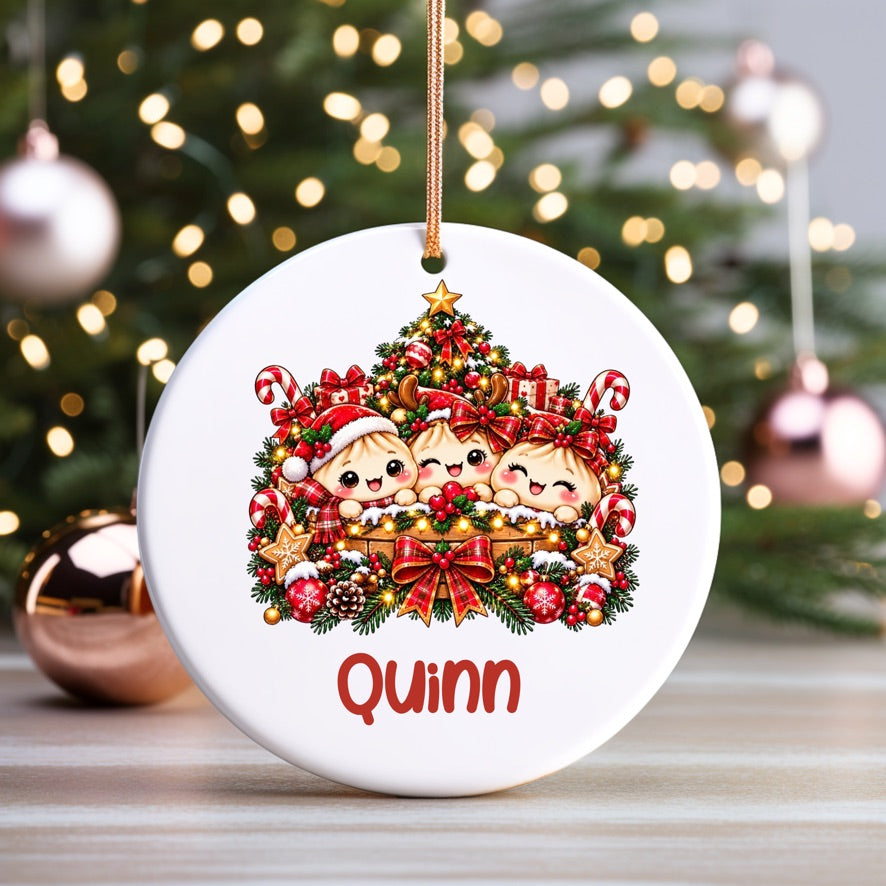 A round white Christmas ornament with a hanging loop, featuring a cartoon illustration of three baby dogs surrounded by a decorated Christmas tree and candy canes, with the name 'Quinn' in red at the bottom. The ornament is shown hanging in front of a blurred Christmas tree with warm lights.