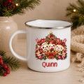 A white enamel-style mug with a silver rim, decorated with a festive Christmas tree graphic featuring three cartoon children and the name 'Quinn' in red text. The mug is styled on a wooden surface with holiday greenery, red berries, and a burlap sack in the background.