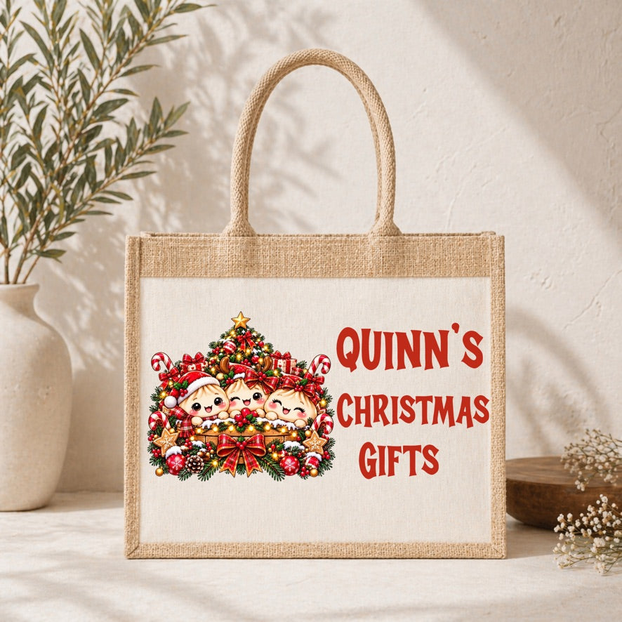 A natural jute tote bag with rope handles, featuring a printed design of three children in a Christmas wreath and the text 'QUINN'S CHRISTMAS GIFTS' in red. The bag is styled on a white surface with a textured wall background, a potted plant, and small dried flowers.