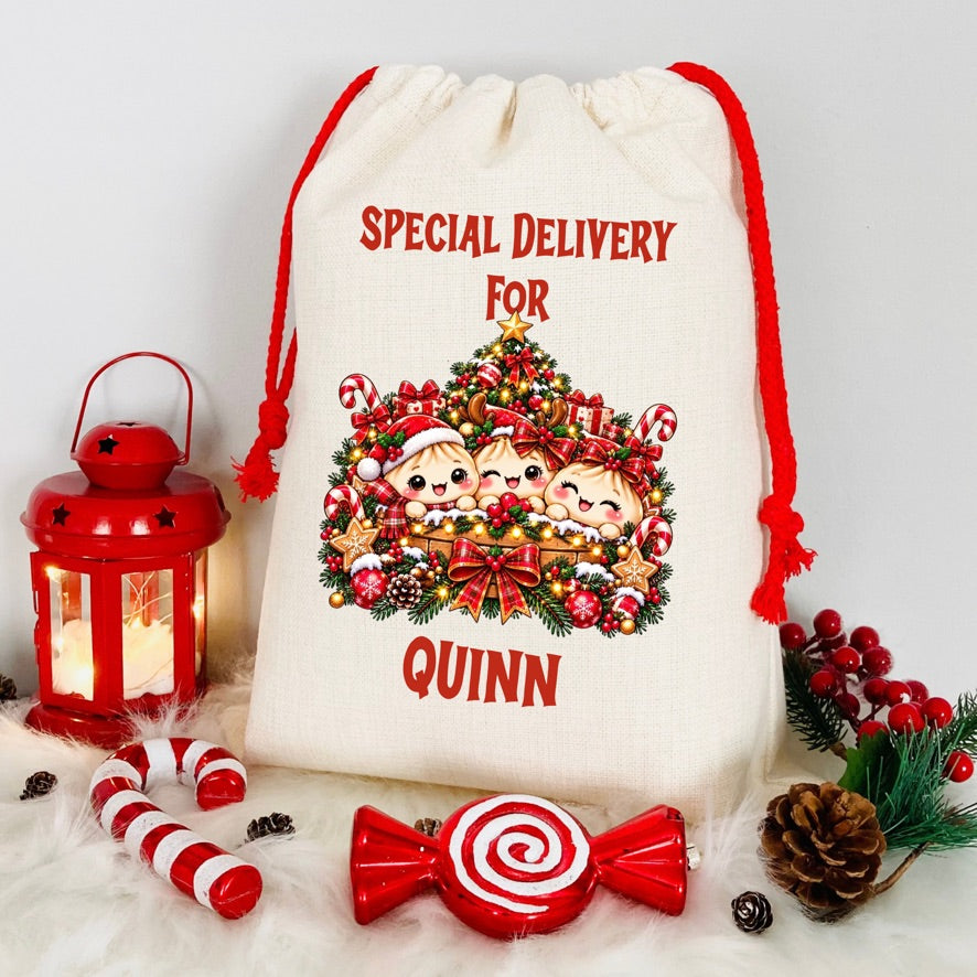 A personalized cream canvas Christmas gift bag with red drawstrings, featuring a festive graphic of three children in Santa hats surrounded by a decorated Christmas tree and the text 'SPECIAL DELIVERY FOR QUINN'. The bag is styled on a white faux fur surface with holiday props including a red lantern, candy canes, a lollipop-shaped ornament, pinecones, and red berries.