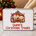 A rectangular metal tin with rounded corners, featuring a white front label with a Christmas tree illustration and the text 'Quinn's Christmas Treats' in red. The tin is photographed on a light wood surface with a blurred Christmas tree and cream fabric in the background.