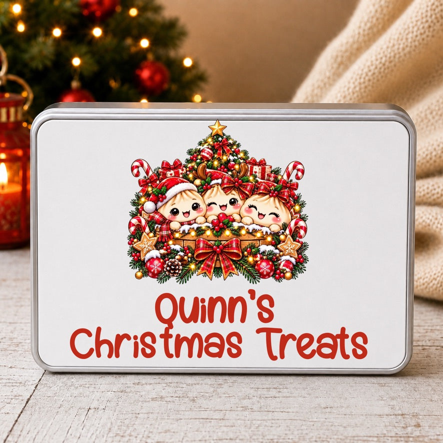 A rectangular metal tin with rounded corners, featuring a white front label with a Christmas tree illustration and the text 'Quinn's Christmas Treats' in red. The tin is photographed on a light wood surface with a blurred Christmas tree and cream fabric in the background.
