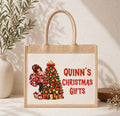 A natural jute tote bag with rope handles, featuring a printed illustration of a girl in a red coat next to a decorated Christmas tree and the red text 'QUINN'S CHRISTMAS GIFTS'. The bag is displayed on a light surface with a textured wall and a potted plant in the background.