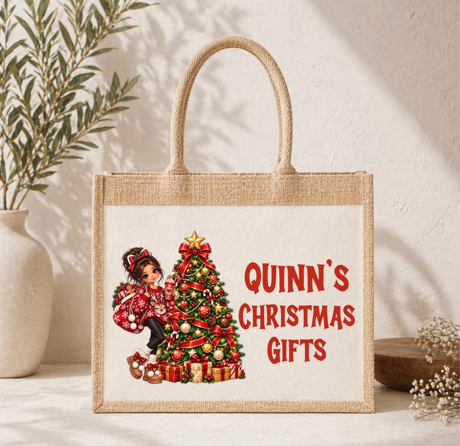A natural jute tote bag with rope handles, featuring a printed illustration of a girl in a red coat next to a decorated Christmas tree and the red text 'QUINN'S CHRISTMAS GIFTS'. The bag is displayed on a light surface with a textured wall and a potted plant in the background.