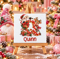 A square Tile print on a small wooden easel, featuring a festive Christmas design with Santa Claus, a reindeer, a large red letter 'Q', gifts, ornaments, and the name 'Quinn' in red script. The scene is decorated with holiday lights, wrapped presents, pinecones, and a gnome figure in the background.