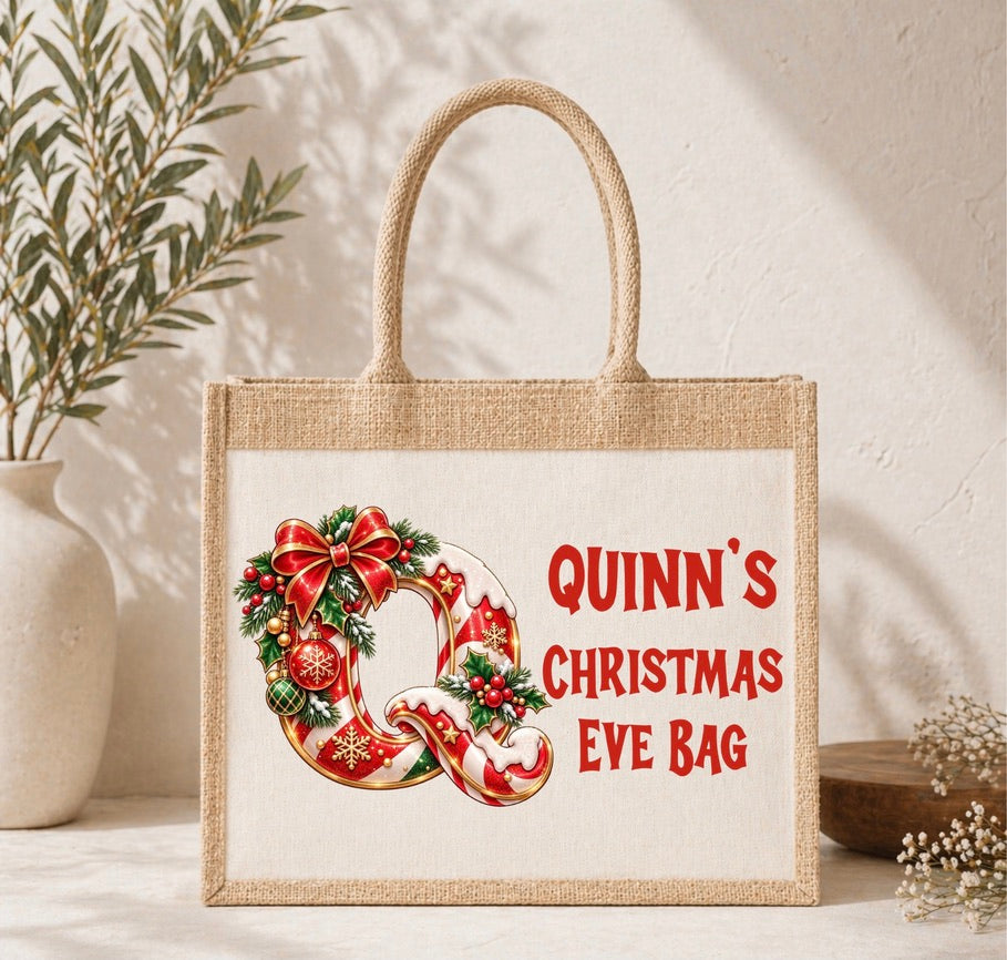 A natural jute tote bag with a white printed panel featuring a red and green Christmas wreath with candy canes and the text 'QUINN'S CHRISTMAS EVE BAG' in red. The bag has two rope handles and is styled on a tabletop with a vase of greenery and a small wooden bowl in the background.