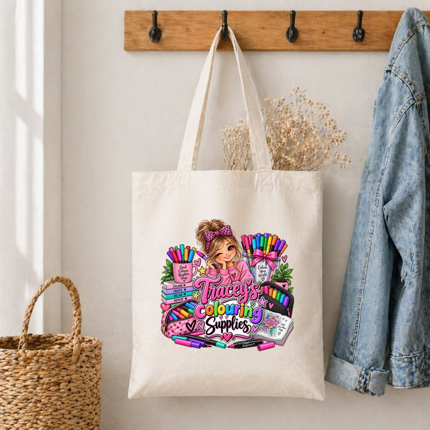 An off-white canvas tote bag hanging on a wooden wall hook, decorated with a colorful graphic of a girl surrounded by crayons and the text 'Tracey's Coloring Supplies'. A blue denim jacket hangs on the right, and a woven basket is on the left.