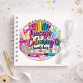 A white spiral-bound notebook with a colorful custom illustration reading 'Tracey's Colouring Swatches' on the cover, styled on a light marble surface with a gold pen, white ribbon, and small confetti.