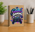 A spiral-bound notebook standing upright on a wooden desk, featuring a colorful gaming-themed cover design with the text 'Quinn's GAMING NOTES' and an illustrated headset and controller. A small potted plant is on the left, and a cup holding pencils is on the right, against a neutral indoor background.
