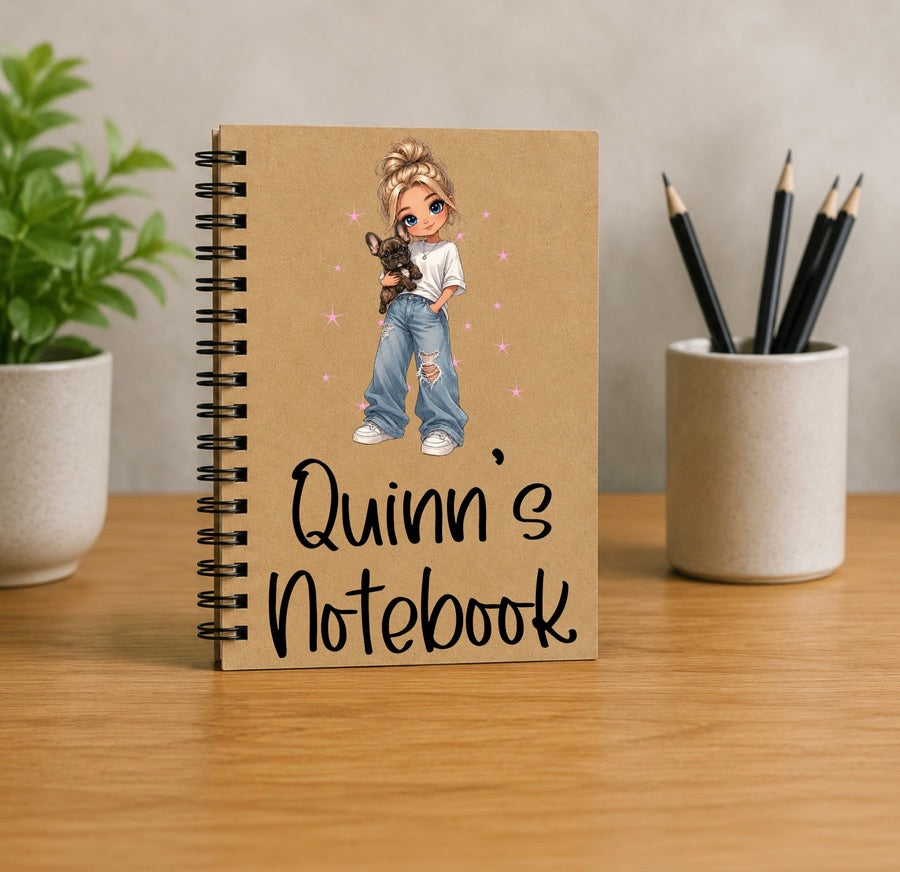 A personalized spiral notebook with a kraft paper cover stands on a wooden desk. The cover features an illustration of a girl with blonde hair in a bun holding a small dog, with the text 'Quinn's Notebook' written in black script.