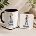 A white ceramic mug with a black handle and interior, alongside a matching square coaster, both printed with a cartoon girl holding a small black cat and the name 'Quinn' in black script. The mug and coaster sit on a beige tabletop with soft-focus decorative plants and a vase in the background.