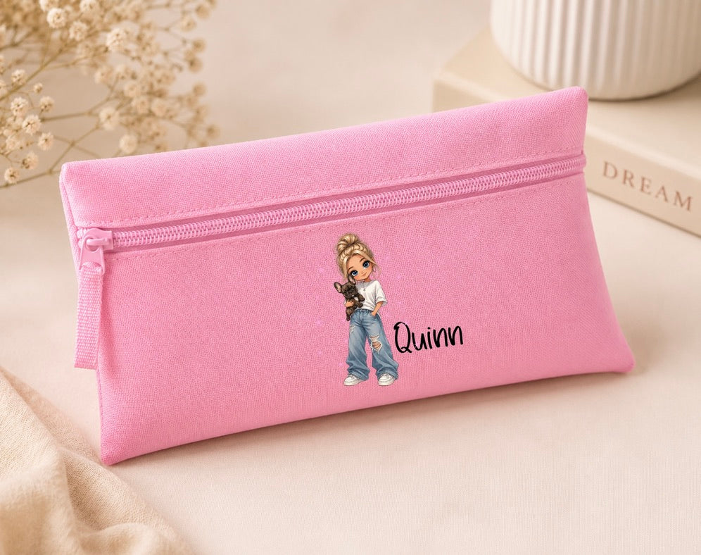 A pink canvas zipper pouch with a cartoon girl illustration and the name 'Quinn' printed on the front, styled on a beige surface with dried flowers and a book in the background.