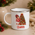 A white enamel-style mug with a silver rim, featuring a Christmas tree illustration and the name 'Quinn' in red text. The mug is styled on a wooden surface with holiday greenery, red berries, and a beige burlap cloth in the background.