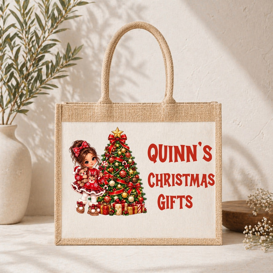 A natural jute tote bag with a printed design featuring a girl in a red dress next to a decorated Christmas tree and the text 'QUINN'S CHRISTMAS GIFTS' in red. The bag is styled on a white surface with a potted plant and dried flowers in the background.