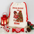 A personalized cream-colored Christmas drawstring gift bag with red tassels, featuring a printed illustration of a girl next to a decorated Christmas tree and the text 'SPECIAL DELIVERY FOR QUINN'. The bag is styled on a white faux fur surface with red holiday props including a lantern, candy canes, a lollipop-shaped ornament, pinecones, and berry sprigs.