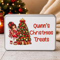 A rectangular tin of Christmas treats with a white lid, featuring an illustration of a girl in a red dress next to a decorated Christmas tree and the text 'Quinn's Christmas Treats' in red. The tin is set on a rustic wooden table with a blurred Christmas tree and cozy background.