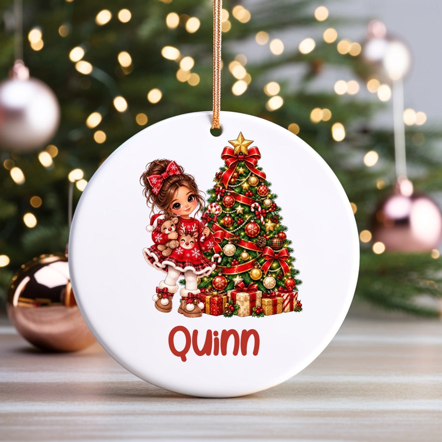 A personalized round white ceramic Christmas ornament hanging in front of a blurred Christmas tree with lights. The ornament features a printed illustration of a young girl in a red dress next to a decorated Christmas tree, with the name 'Quinn' written in red at the bottom.