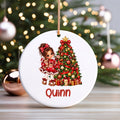 A personalized round white ceramic Christmas ornament hanging in front of a blurred Christmas tree with lights. The ornament features a printed illustration of a young girl in a red dress next to a decorated Christmas tree, with the name 'Quinn' written in red at the bottom.
