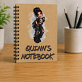 A spiral-bound notebook standing upright on a wooden desk. The notebook cover is tan with a black metal spiral and features an illustration of a punk-style girl with the text 'QUINN'S NOTEBOOK' in black.