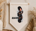 A personalized canvas wall banner featuring a cartoon girl with messy black hair, a skull top, and ripped jeans, with the name 'QUINN' printed in black. The banner is displayed hanging on a beige wall with boho decor props like a macrame curtain and dried flowers.