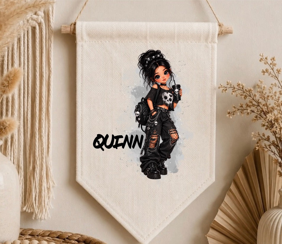 A personalized canvas wall banner featuring a cartoon girl with messy black hair, a skull top, and ripped jeans, with the name 'QUINN' printed in black. The banner is displayed hanging on a beige wall with boho decor props like a macrame curtain and dried flowers.