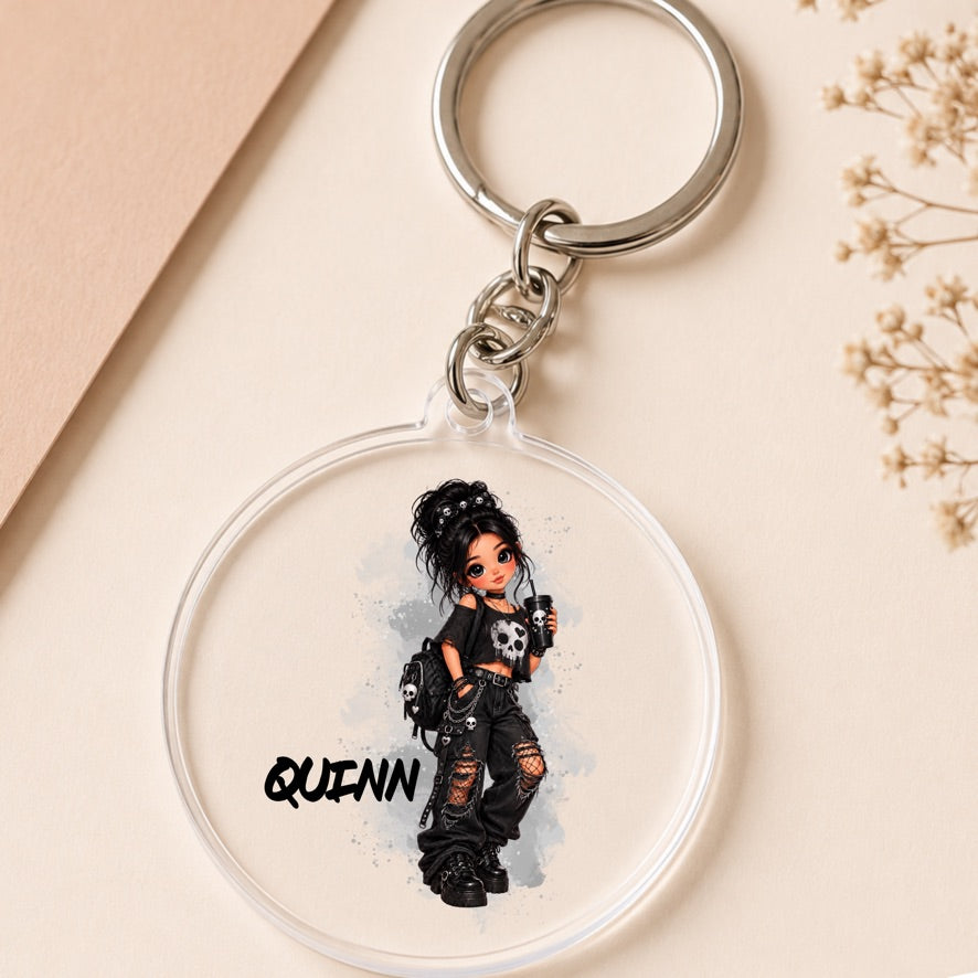 A round clear acrylic keychain featuring a cartoon illustration of a girl with messy dark hair, a black top, ripped jeans, and a skull, with the name 'QUINN' printed below. The keychain is shown on a beige background with dried flowers and a tan paper corner.