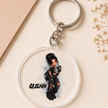 A round clear acrylic keychain featuring a cartoon illustration of a girl with messy dark hair, a black top, ripped jeans, and a skull, with the name 'QUINN' printed below. The keychain is shown on a beige background with dried flowers and a tan paper corner.