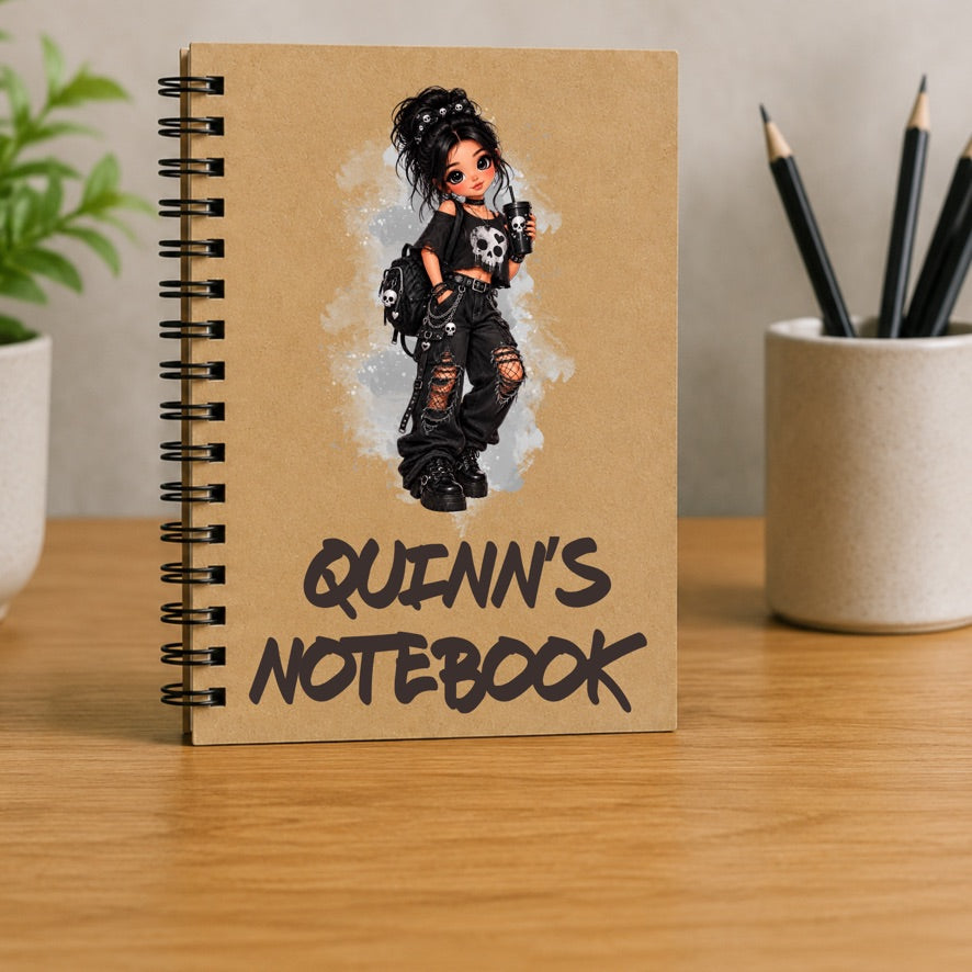 A spiral-bound notebook standing upright on a wooden desk. The notebook cover is tan with a black metal spiral and features an illustration of a punk-style girl with the text 'QUINN'S NOTEBOOK' in black.