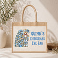 A personalized jute tote bag with rope handles, featuring a printed design of a blue and gold Christmas nutcracker next to a decorated tree and the text 'QUINN'S CHRISTMAS EVE BAG' in blue. The bag is styled on a light beige textured surface with a potted olive branch and small flowers in the background.