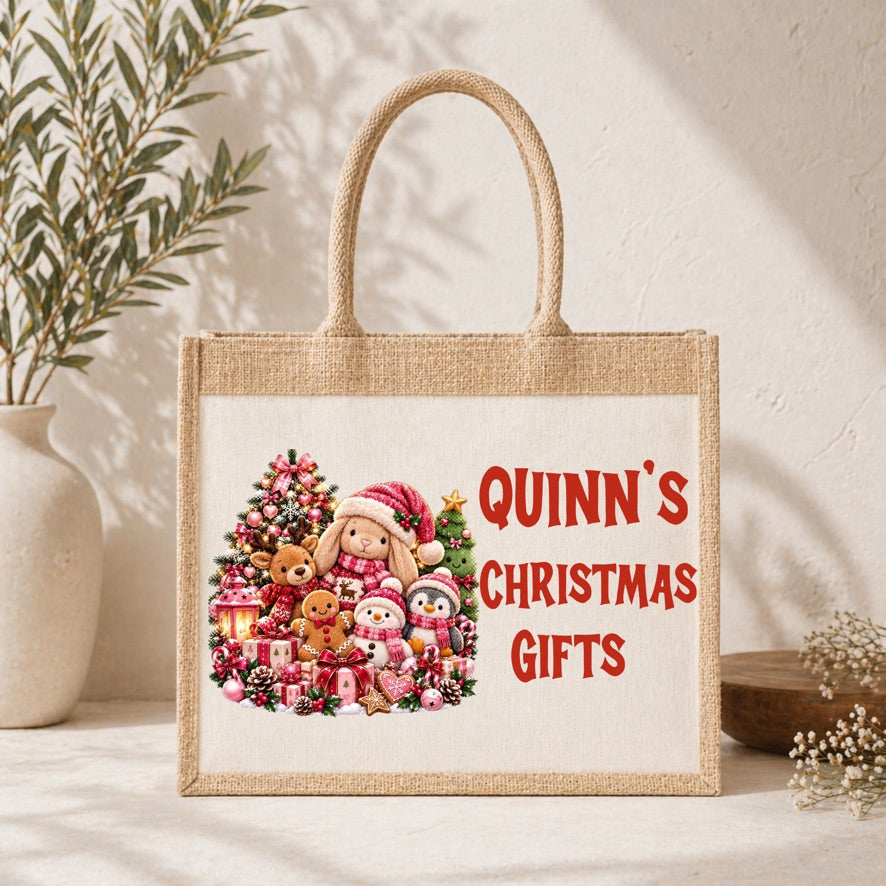 A natural jute tote bag with rope handles stands on a light surface against a textured wall. The front has a printed holiday illustration of a bunny, teddy bear, snowman, and Christmas tree alongside red text reading “QUINN'S CHRISTMAS GIFTS.