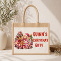 A natural jute tote bag with rope handles stands on a light surface against a textured wall. The front has a printed holiday illustration of a bunny, teddy bear, snowman, and Christmas tree alongside red text reading “QUINN'S CHRISTMAS GIFTS.