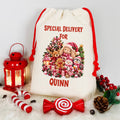 A cream canvas Christmas gift bag with red drawstrings, printed with 'SPECIAL DELIVERY FOR QUINN' and a festive teddy bear and snowman illustration. The bag is styled on a white faux-fur surface with red holiday props including a lantern, candy canes, a lollipop-shaped ornament, pinecones, and berry sprigs.