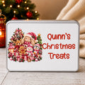 A rectangular tin of Christmas treats with a white lid, featuring a festive illustration of teddy bears, a bunny, snowman, and penguin next to the red text 'Quinn's Christmas Treats'. The tin is set on a wooden surface with a blurred Christmas tree and cozy background.
