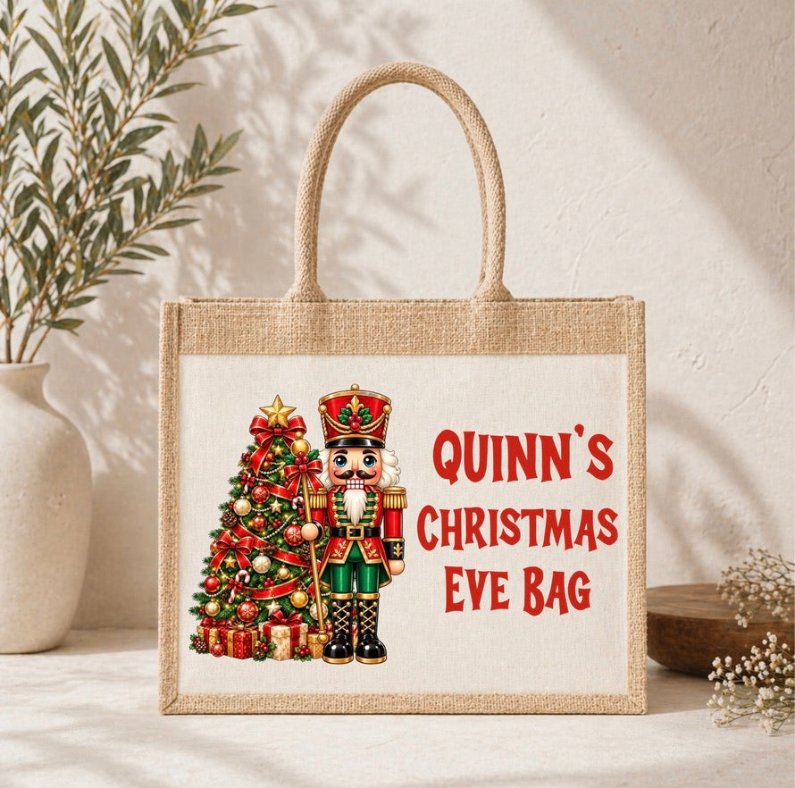 A personalized jute tote bag with a Christmas nutcracker and tree design, featuring the text 'QUINN'S CHRISTMAS EVE BAG' in red, set against a warm neutral background with decorative plants.