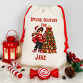 A cream-colored canvas Christmas gift bag with red drawstrings, featuring a printed illustration of a child in a Santa hat next to a decorated Christmas tree, with the text 'SPECIAL DELIVERY FOR JAKE'. The bag is styled on a white faux fur surface with red holiday props including a lantern, candy canes, a lollipop, pinecones, and berry sprigs.