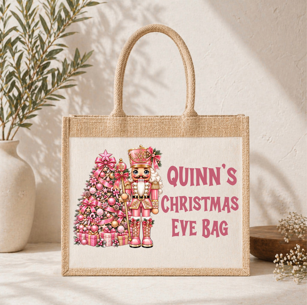 A natural jute tote bag with a printed Christmas nutcracker and pink tree design, featuring the text 'QUINN'S CHRISTMAS EVE BAG' in pink, set against a warm neutral background with decorative plants.
