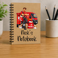 A spiral-bound notebook standing upright on a wooden desk. The notebook cover is kraft brown and features an illustration of a postal worker in a red uniform standing beside a red van and mail bags, with the text 'Nick's Notebook' written in black script.