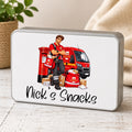 A rectangular metal tin with rounded corners and a silver-gray edge, featuring a printed illustration of a postman standing beside a red van and post box, with the text 'Nick's Snacks' below. The tin is photographed on a light wood surface with a soft neutral fabric and potted plant in the background.