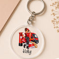 A personalized acrylic keychain featuring an illustration of a female postal worker in red next to a red post box, van, and packages, with the name 'Kelly' printed below. The keychain is shown on a beige background with a kraft paper corner and dried flowers.