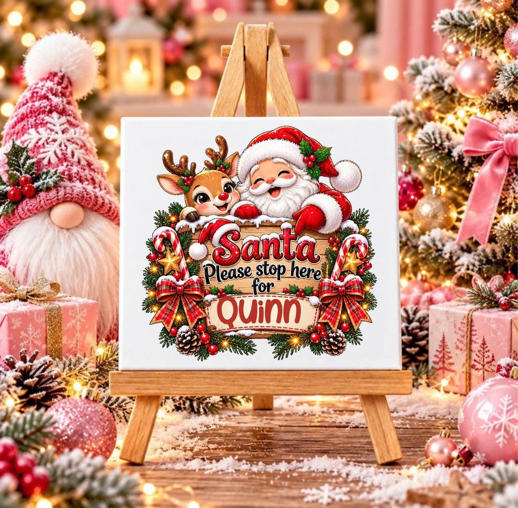 A square tile on a small wooden easel, featuring a festive Christmas design with Santa Claus, a reindeer, candy canes, pinecones, and the personalized text 'Santa Please stop here for Quinn'. The scene is decorated for Christmas with a gnome, wrapped gifts, and a lit Christmas tree in the background.