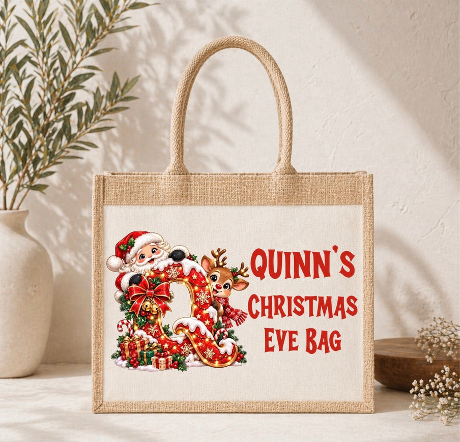 A personalized jute tote bag with a Christmas Eve theme, featuring a cartoon Santa and reindeer next to red text that reads 'QUINN'S CHRISTMAS EVE BAG'. The bag is styled on a light surface with a vase and dried flowers in the background.