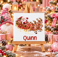 A square canvas print on a small wooden easel, featuring a festive Christmas design with Santa Claus in a red sleigh pulled by three golden-brown reindeer, and the name 'Quinn' in red script below. The canvas is styled in a cozy, blurred Christmas setting with a gnome, wrapped gifts, and a decorated tree in the background.