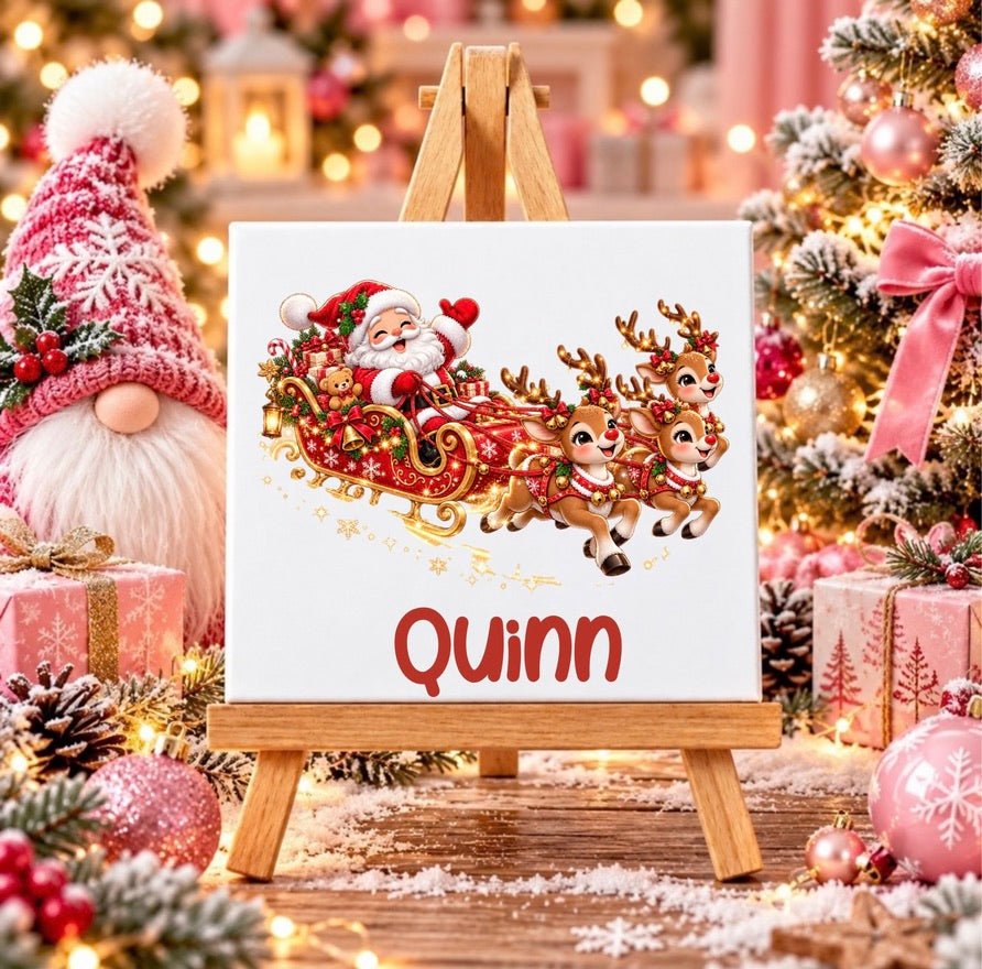 A square canvas print on a small wooden easel, featuring a festive Christmas design with Santa Claus in a red sleigh pulled by three golden-brown reindeer, and the name 'Quinn' in red script below. The canvas is styled in a cozy, blurred Christmas setting with a gnome, wrapped gifts, and a decorated tree in the background.