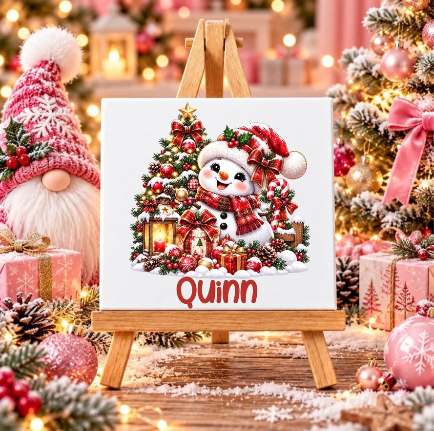 Personalised snowman Christmas ceramic tile with custom name, featuring a festive snowman, Christmas tree and presents displayed on a wooden stand.