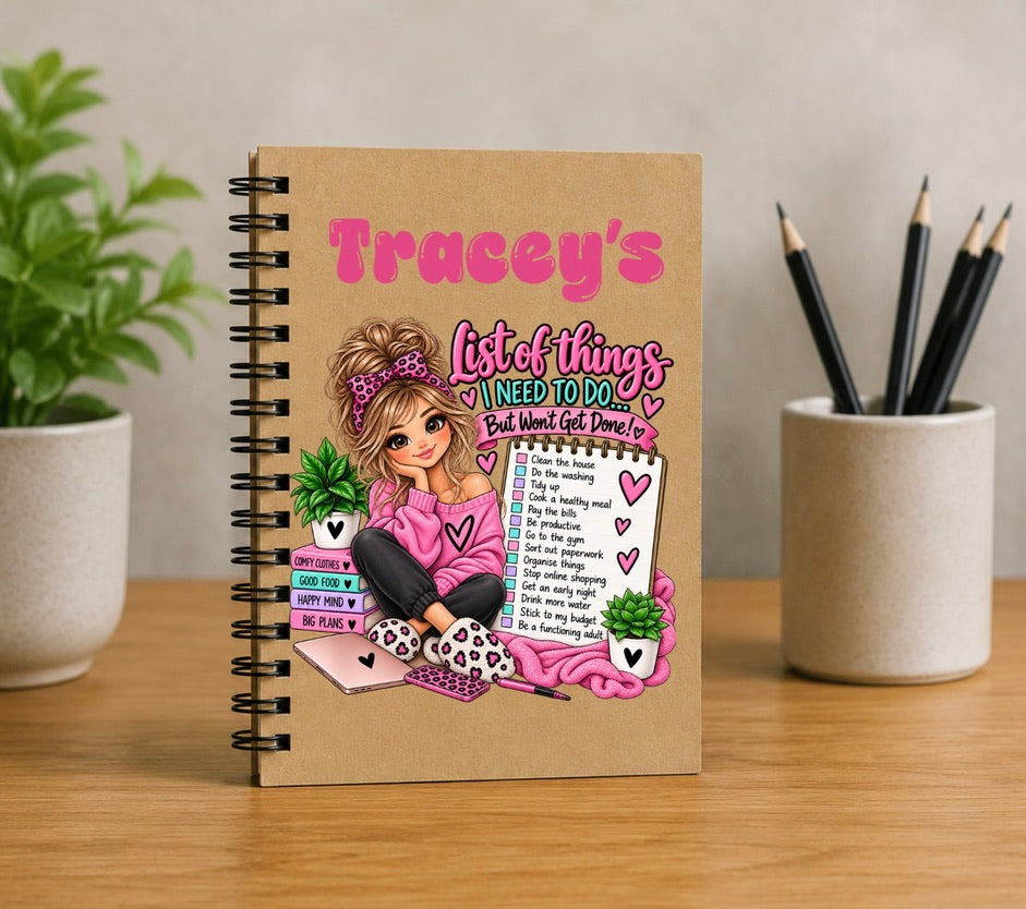 A spiral-bound notebook with a kraft paper cover stands on a wooden table. The cover features a colorful cartoon illustration of a woman with the title 'Tracey's List of things I need to do' in pink and teal lettering.