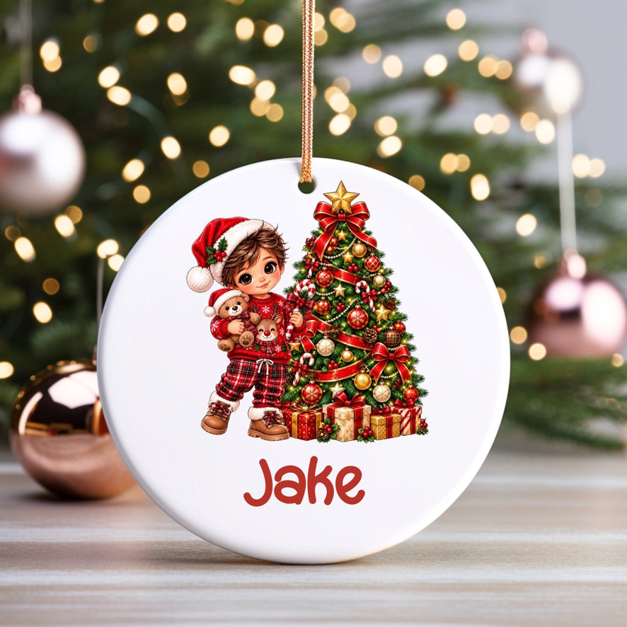A personalized round white Christmas ornament featuring an illustration of a young boy in a Santa hat hugging a decorated Christmas tree, with the name 'Jake' printed below in red. The ornament hangs from a gold string against a blurred festive background with a lit Christmas tree and reflective ornaments.