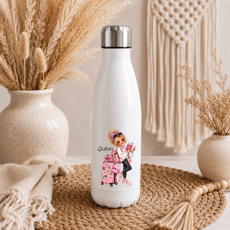 A white insulated water bottle with a silver cap stands on a woven mat in a styled indoor setting. The bottle has a printed illustration of a woman with a pink suitcase and the word 'Queen' on the front.