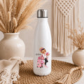 A white insulated water bottle with a silver cap stands on a woven mat in a styled indoor setting. The bottle has a printed illustration of a woman with a pink suitcase and the word 'Queen' on the front.